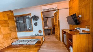 Una habitación moderna con suelo de madera, cama con cabecero, televisor, escritorio con silla y baño visible a través de una puerta abierta.