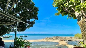 Vista de una playa con una terraza de madera, plantas en macetas y sillas rojas. Se ven barcos en el agua.
