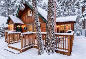 En stuga av trä med en veranda och en öppen spis, omgiven av snötäckta träd.