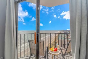 Un balcón con vistas despejadas a la playa y al paisaje urbano, una silla, una mesita con frutas y bebidas, y una ventana con cortinas.