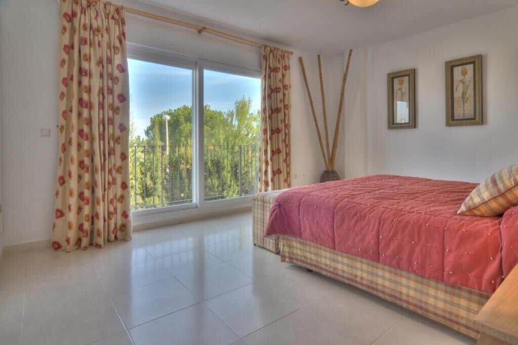 Une chambre avec une grande fenêtre, un lit recouvert d’une courtepointe rouge et deux tableaux encadrés accrochés au mur.
