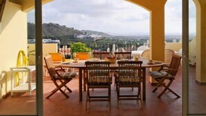 A dining area with a table and chairs, a view of a hillside, and a yellow chair on the left.