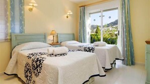 A hotel room with two beds, a window with curtains, and a view of greenery outside.