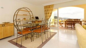 A modern living room with a glass dining table, orange chairs, a sofa, and a view of the city through a large window.