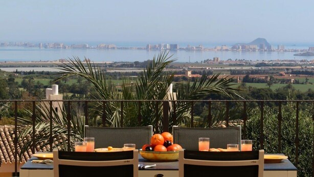 Una mesa de comedor con sillas y un frutero, con vistas a una ciudad costera y una montaña distante.