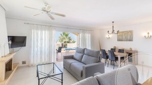 Una sala de estar moderna con un sofá, una mesa de centro de vidrio, un comedor con mesa y sillas, un televisor en la pared y un ventilador de techo.