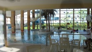 An indoor swimming pool with a view of a tropical garden, surrounded by white plastic chairs and a glass railing. - Ferienapartment-ruegen (Rügen)