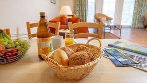 A breakfast table with bread, fruit, juice, and a bottle. There is a wicker basket, a lamp, and a chair in the background. - Ferienapartment-ruegen (Rügen)
