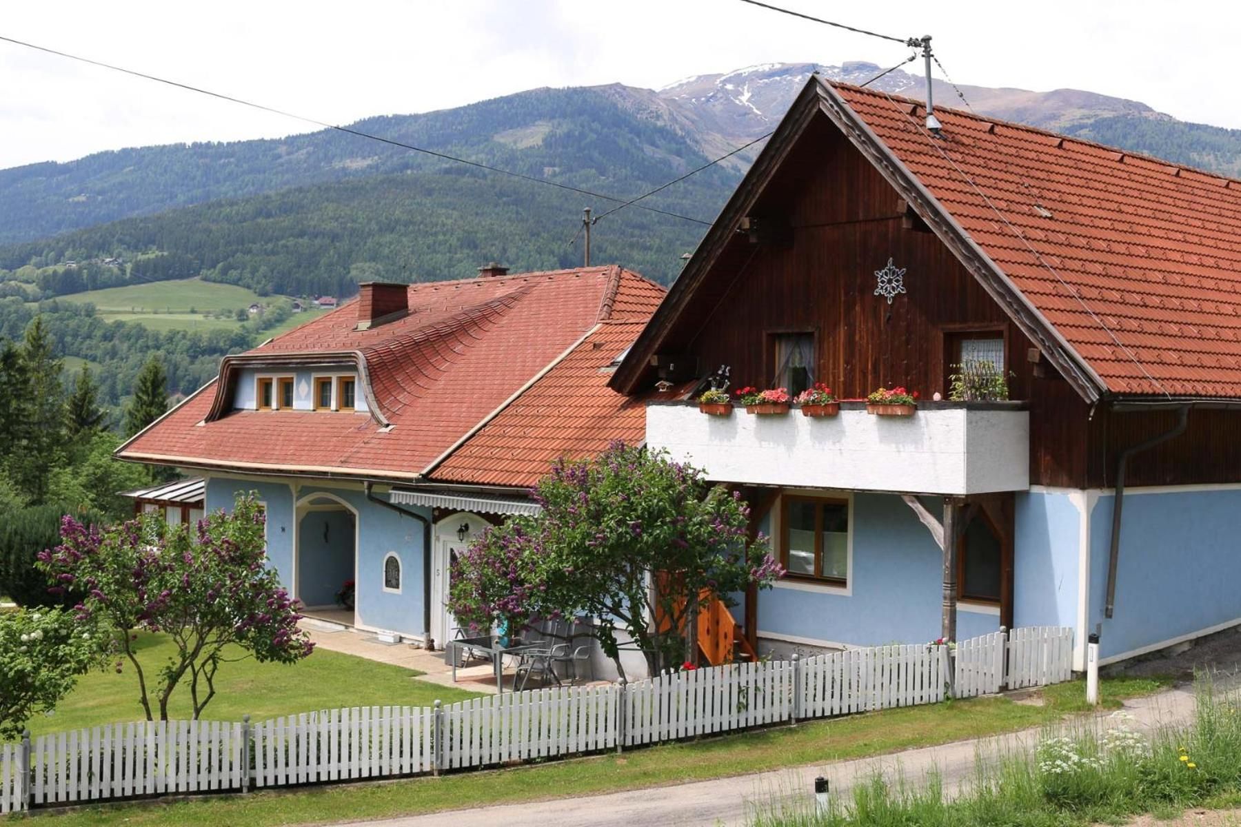 A traditional wooden house with a sloped roof, a balcony with potted plants, and a white picket fence.