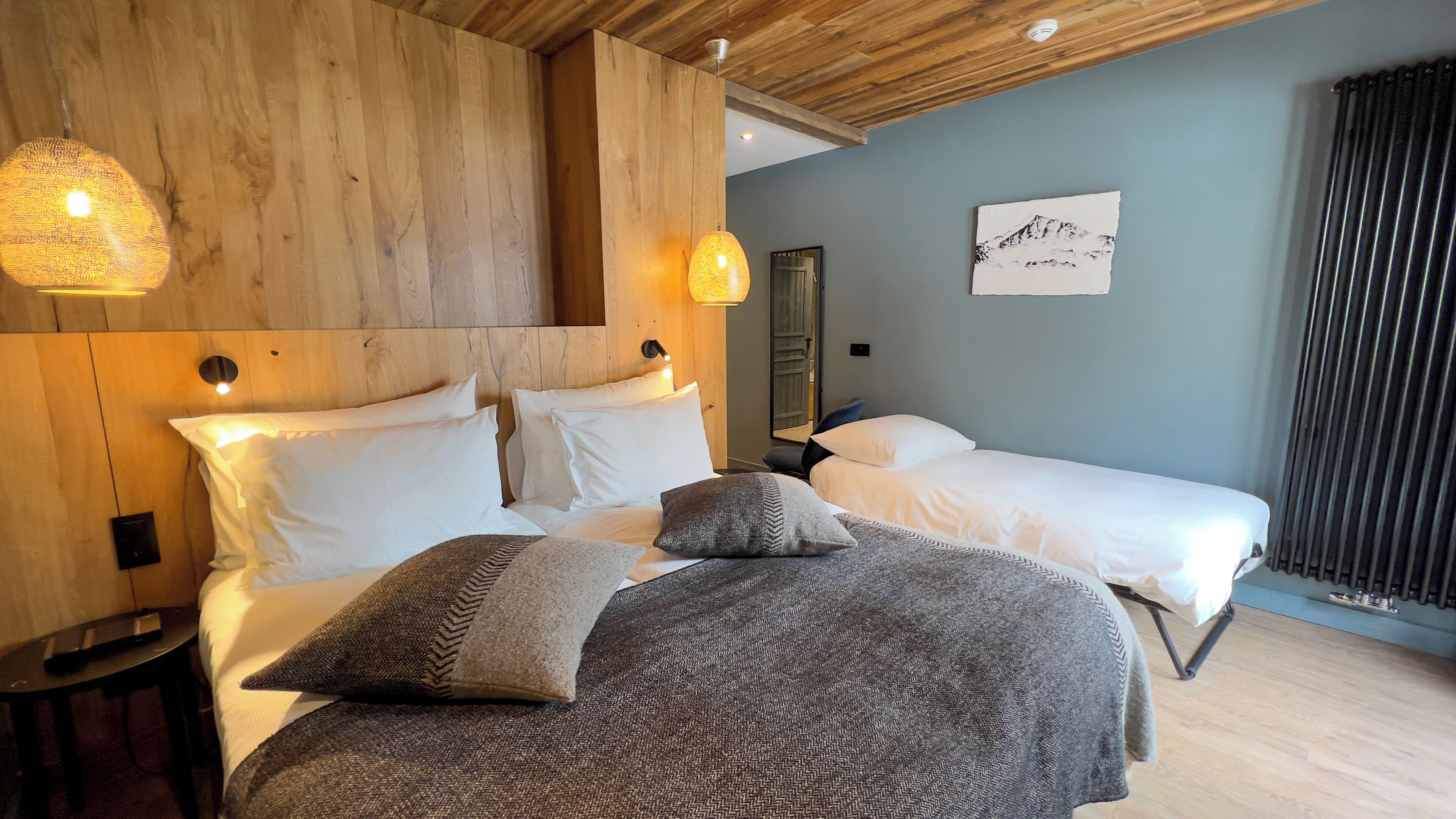 Une chambre d’hôtel avec un plafond en bois, deux lits avec une literie blanche, un plaid gris à texture et une œuvre d’art accrochée au mur.