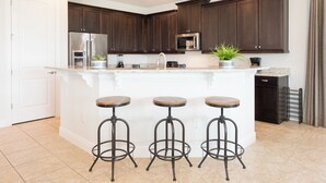 A kitchen with dark wood cabinets, a stainless steel refrigerator, a microwave, a sink, and three bar stools. - Private Pool, Game Room & Waterpark Access – 141LA (Reunion)