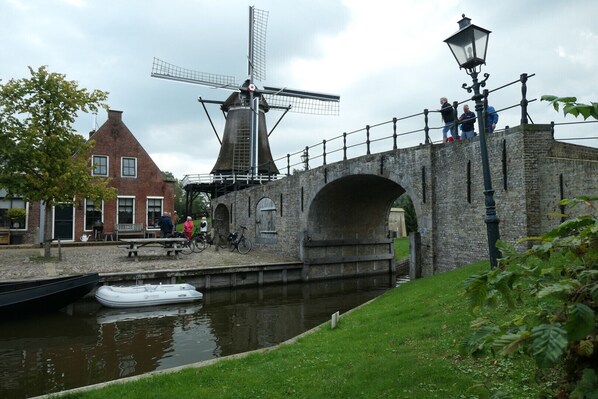 Un pont historique avec un moulin à vent en arrière-plan, un canal avec des bateaux et une zone herbeuse où se trouvent des personnes.