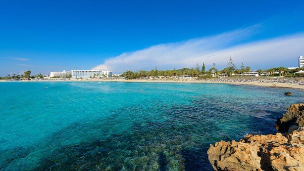 A coastal hotel with clear turquoise waters, rocky shoreline, and a beach with umbrellas.