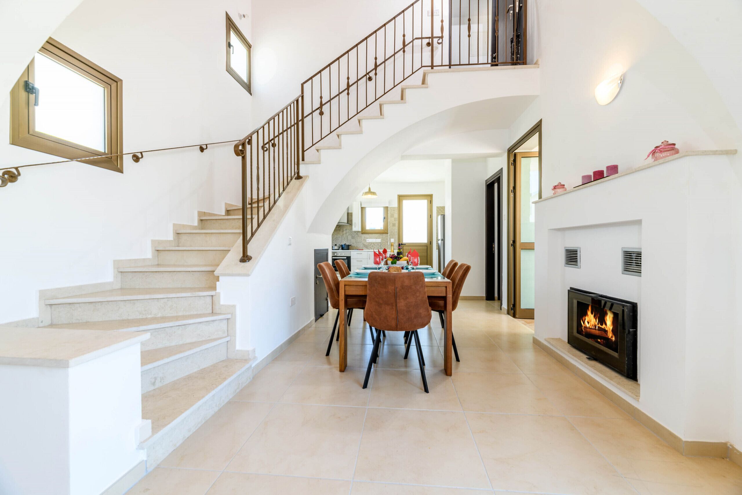 A spacious living area with a staircase, a dining table with chairs, a fireplace, and a kitchen in the background.