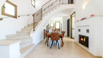 A spacious living area with a staircase, a dining table with chairs, a fireplace, and a kitchen in the background.