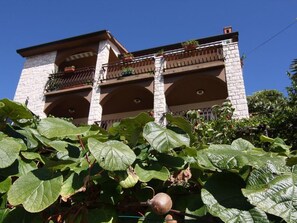 A building with a stone facade, multiple balconies with potted plants, and large green leaves in the foreground. - Family-friendly apartment with terrace and wonderful sea view (Rabac)