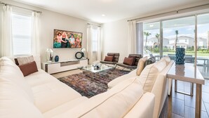 A modern living room with a large sofa, a glass coffee table, and a view of the outdoors. - Home near Disney: Air Hockey Table, Waterpark & Resort Access - 241BU (Reunion)