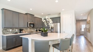 A modern kitchen with grey cabinets, a large island with bar stools, a microwave, and a refrigerator. - Home near Disney: Air Hockey Table, Waterpark & Resort Access - 241BU (Reunion)