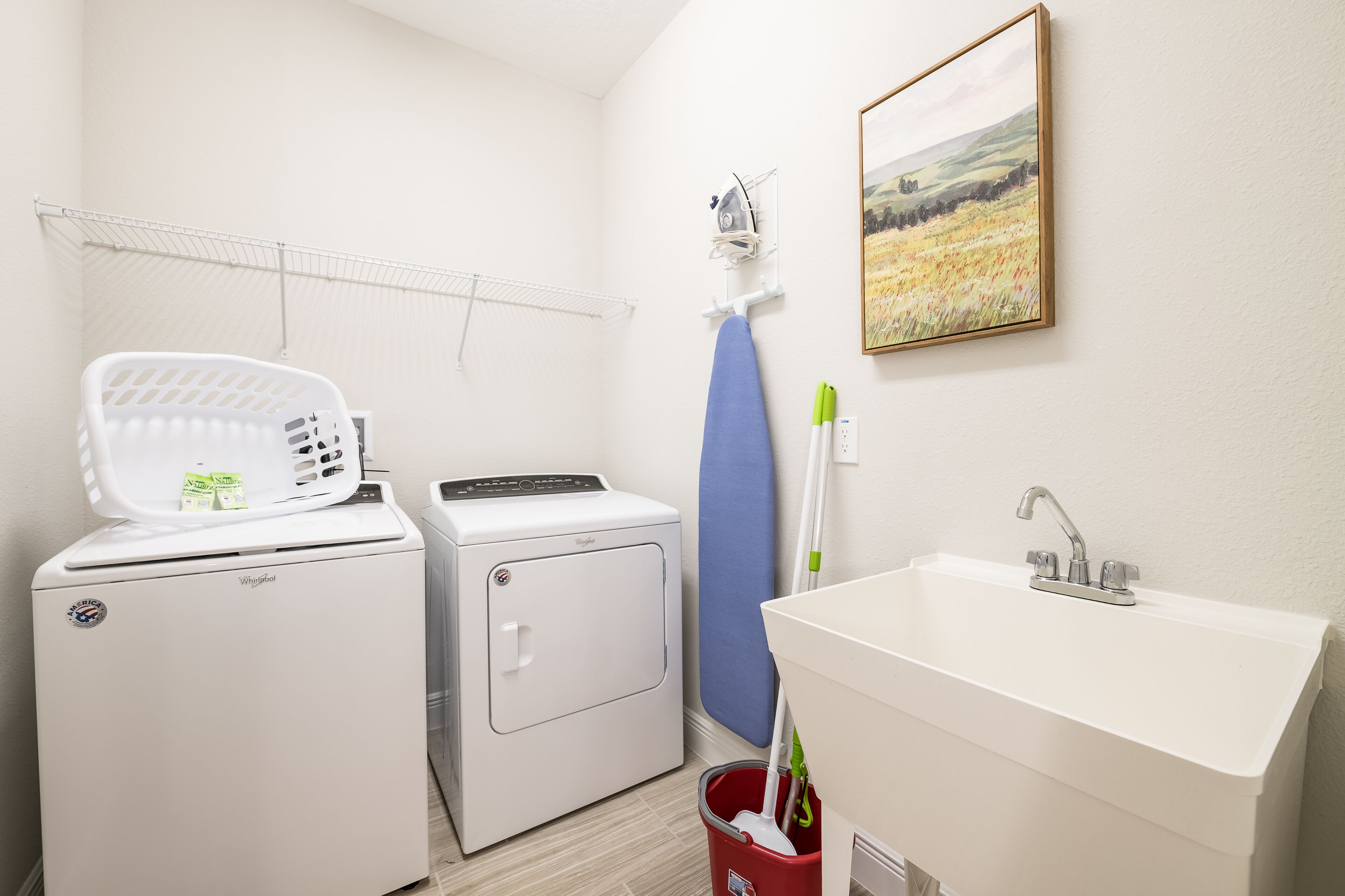 A laundry area with a white washer and dryer, a drying rack, a painting of a field, and a sink with a faucet.