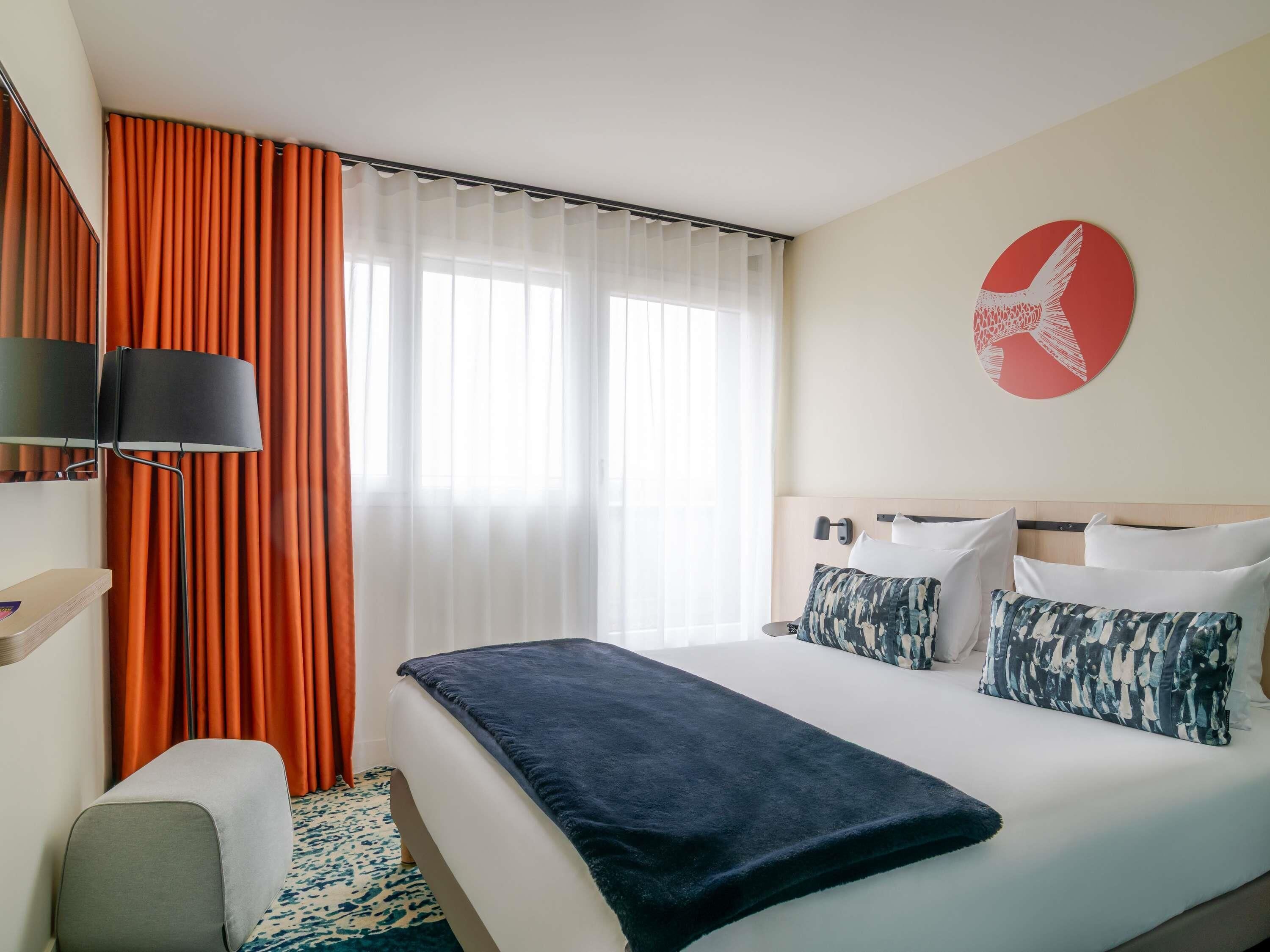 Habitació d'hotel amb llit, escriptori, cadira, làmpada, cortines i una obra d'art a la paret.