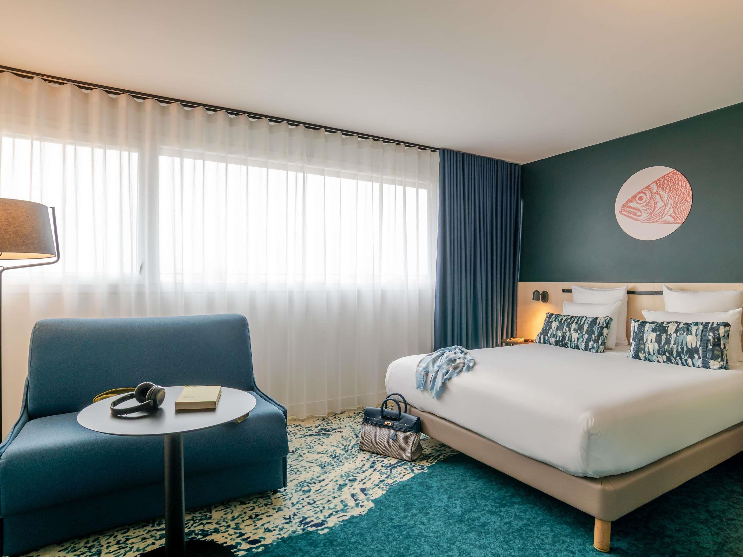 A hotel room with a bed, a blue armchair, a small round table, a lamp, and a wall art piece.