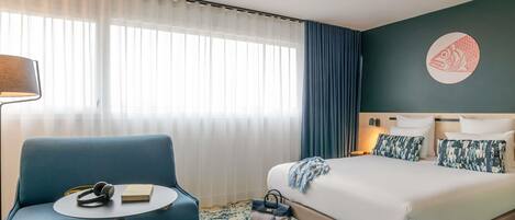 A hotel room with a bed, a blue armchair, a small round table, a lamp, and a wall art piece.