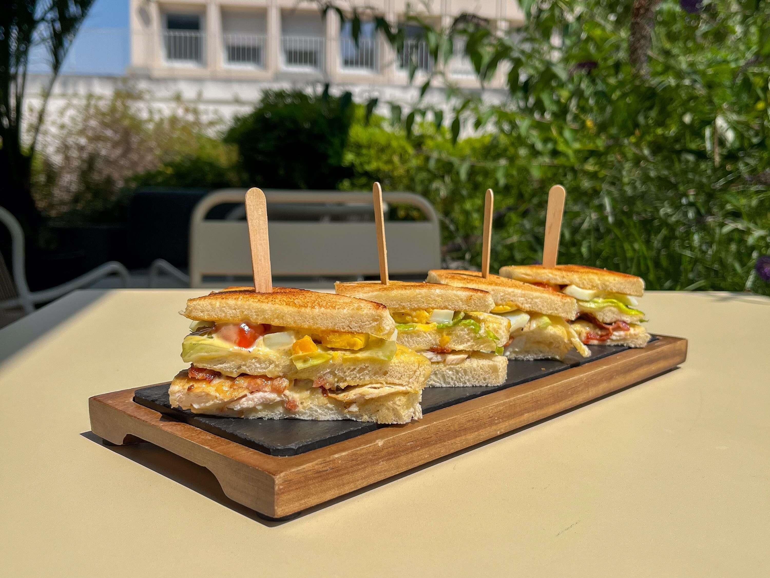 A wooden serving board with four sandwiches, each held together by wooden skewers.
