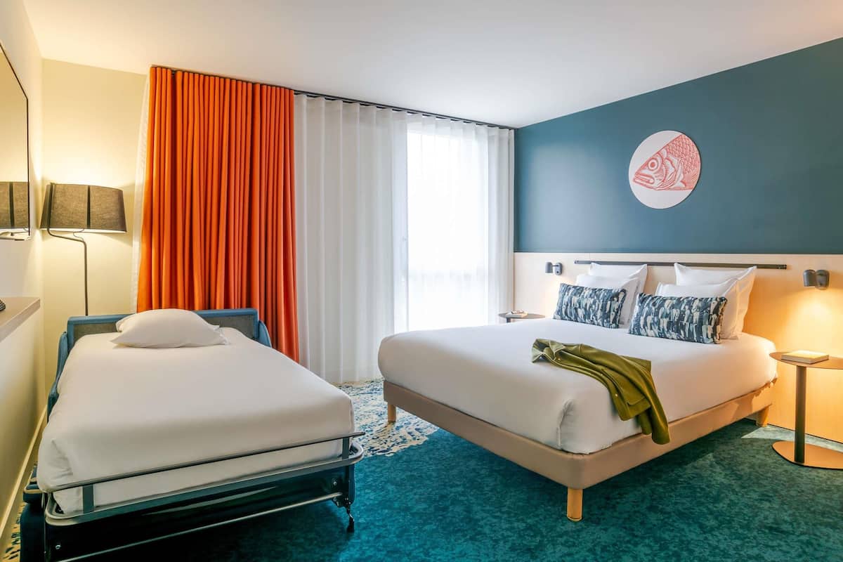 A hotel room with two beds, a large window with curtains, a wall-mounted artwork, and a blue carpet.