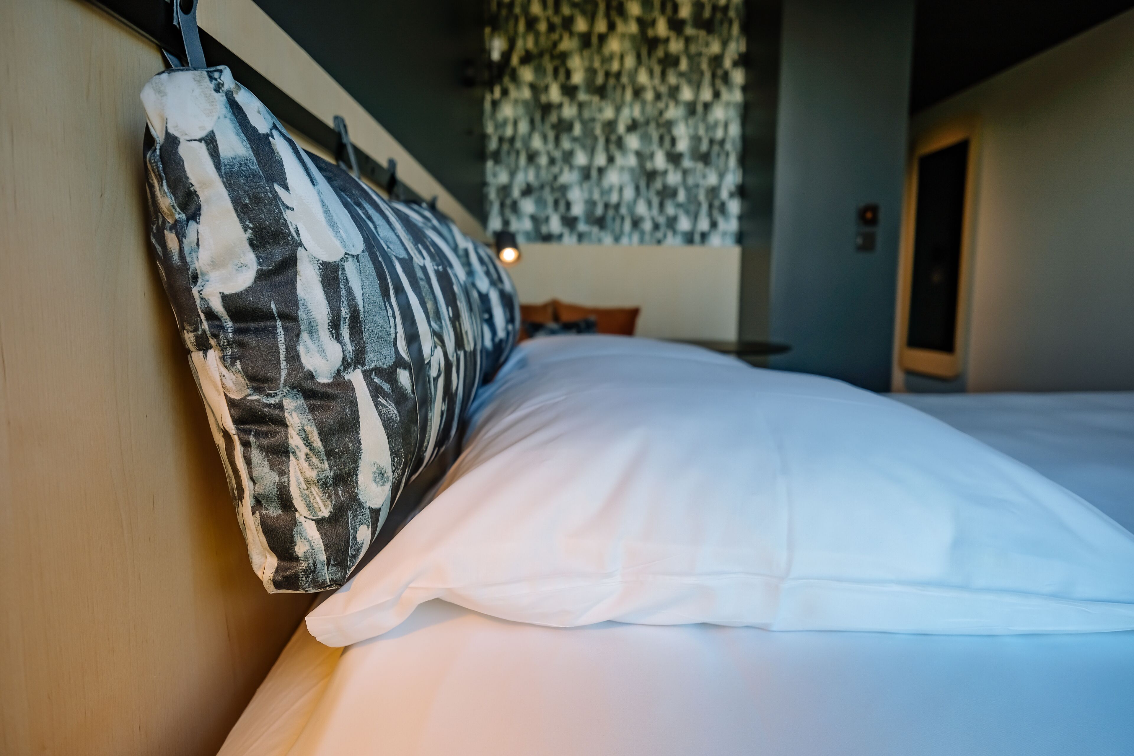 A bed with a patterned pillow and white sheets, a hanging light fixture, and a door in the background.