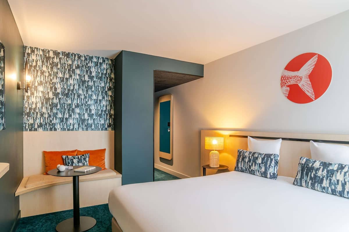 A hotel room with a bed, a seating area with a table, a wall-mounted lamp, and a decorative wall piece.