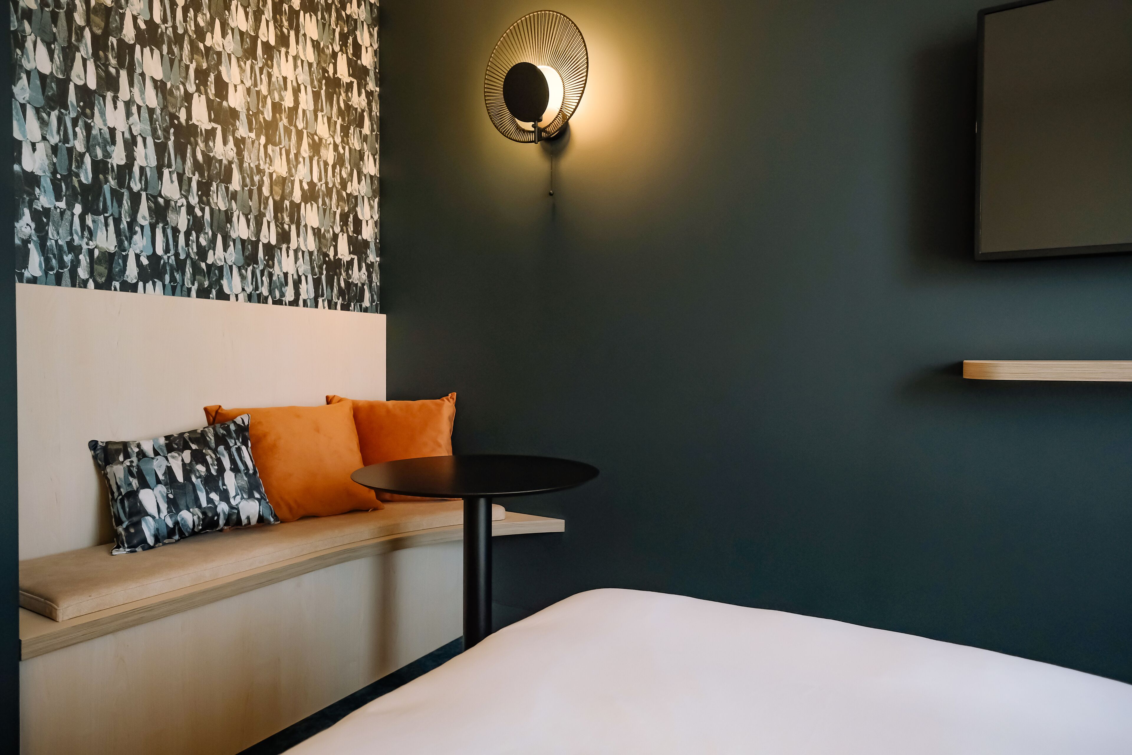 Ein modernes Hotelzimmer mit dunkler Wand, einer runden Wandleuchte, einem Flachbildfernseher, einer Holzbank mit orangefarbenen Kissen und einem kleinen runden Tisch.