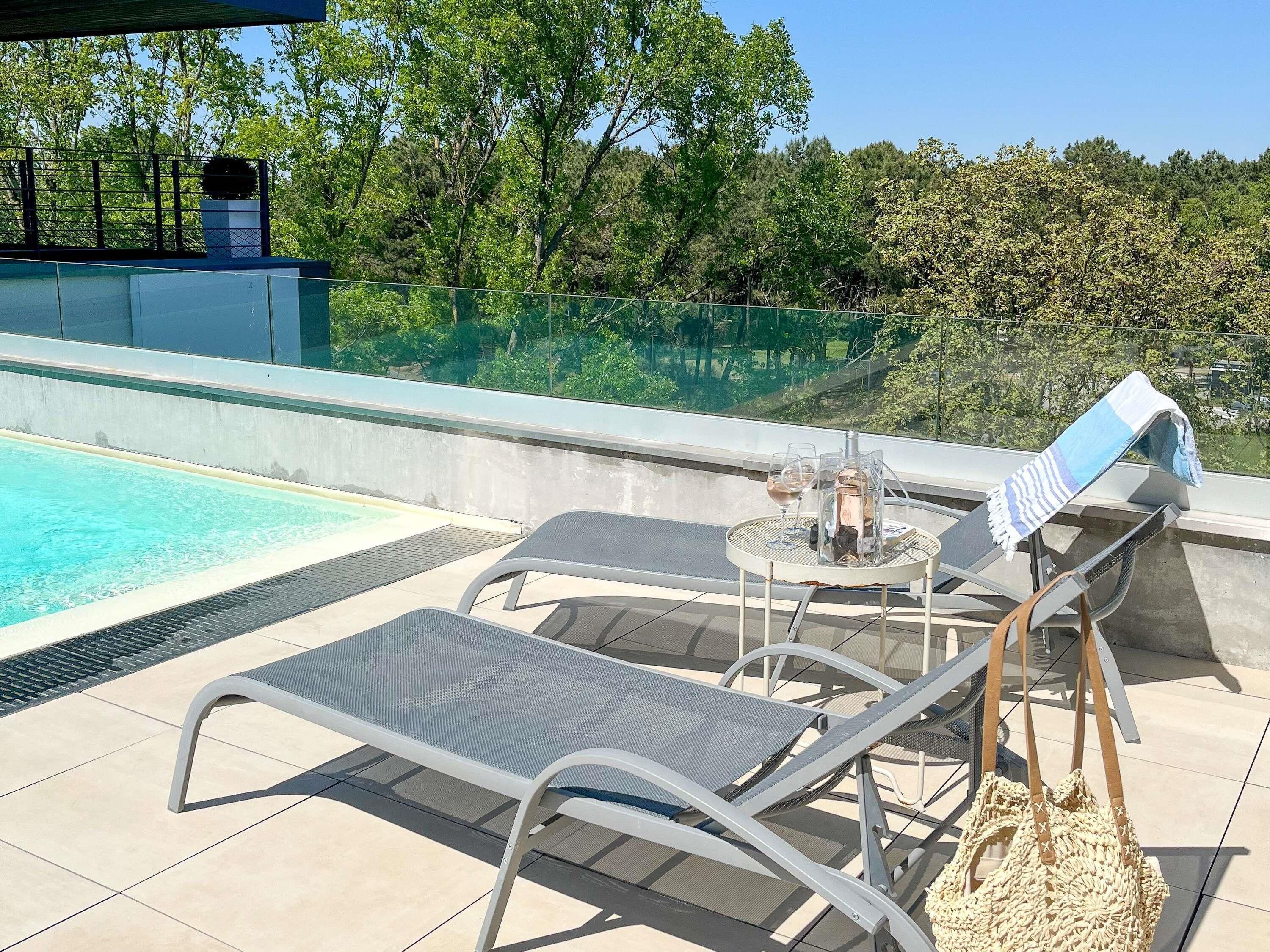 Ein Poolbereich mit Liegestühlen, einem Tisch mit Flasche und Gläsern sowie Blick auf Bäume und ein Gebäude im Hintergrund.