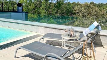 A poolside area with lounge chairs, a table with a bottle and glasses, and a view of trees and a building in the background.