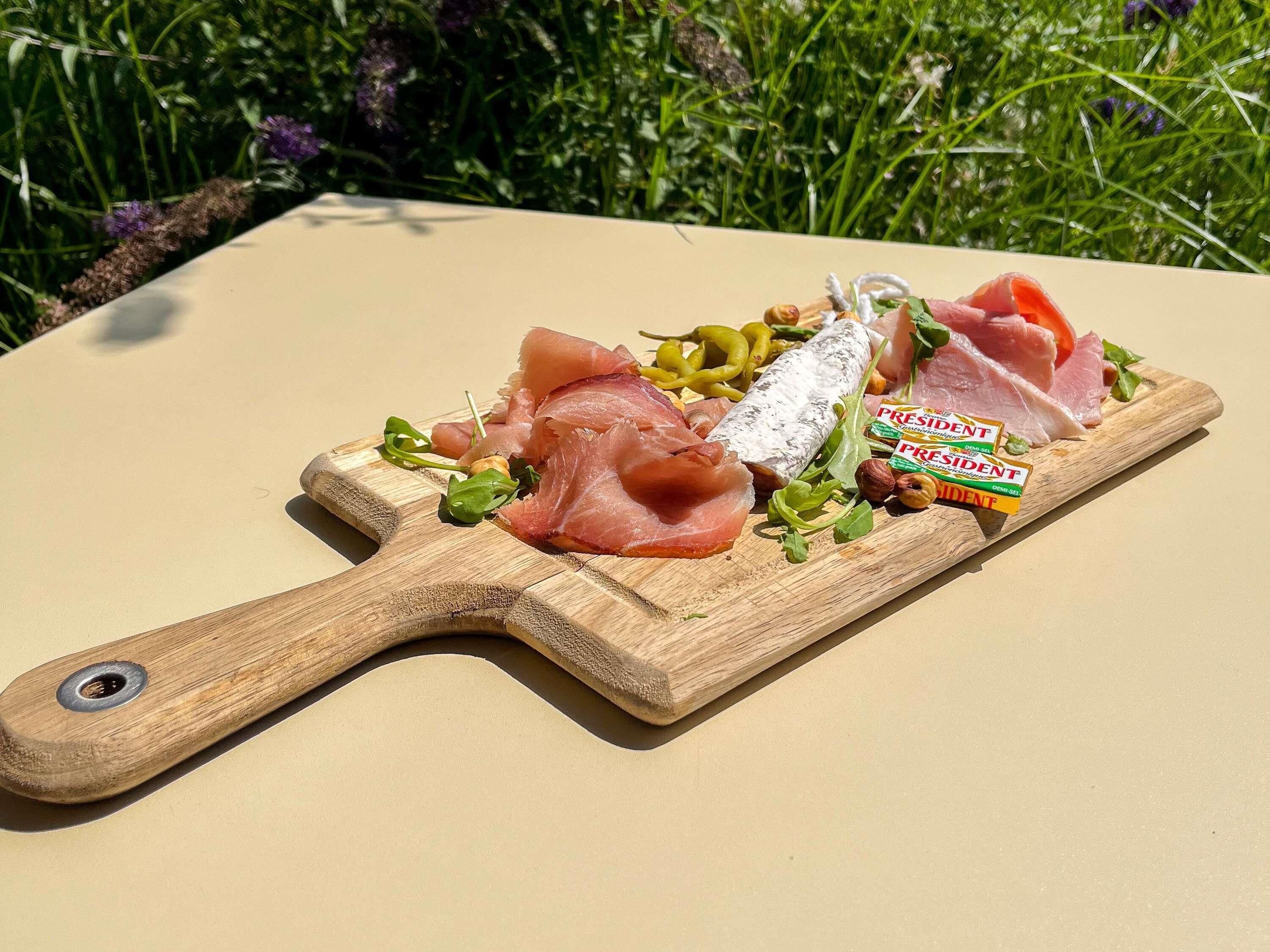 Een houten plank met verschillende vleessoorten en groenten, op een beige oppervlak buiten.