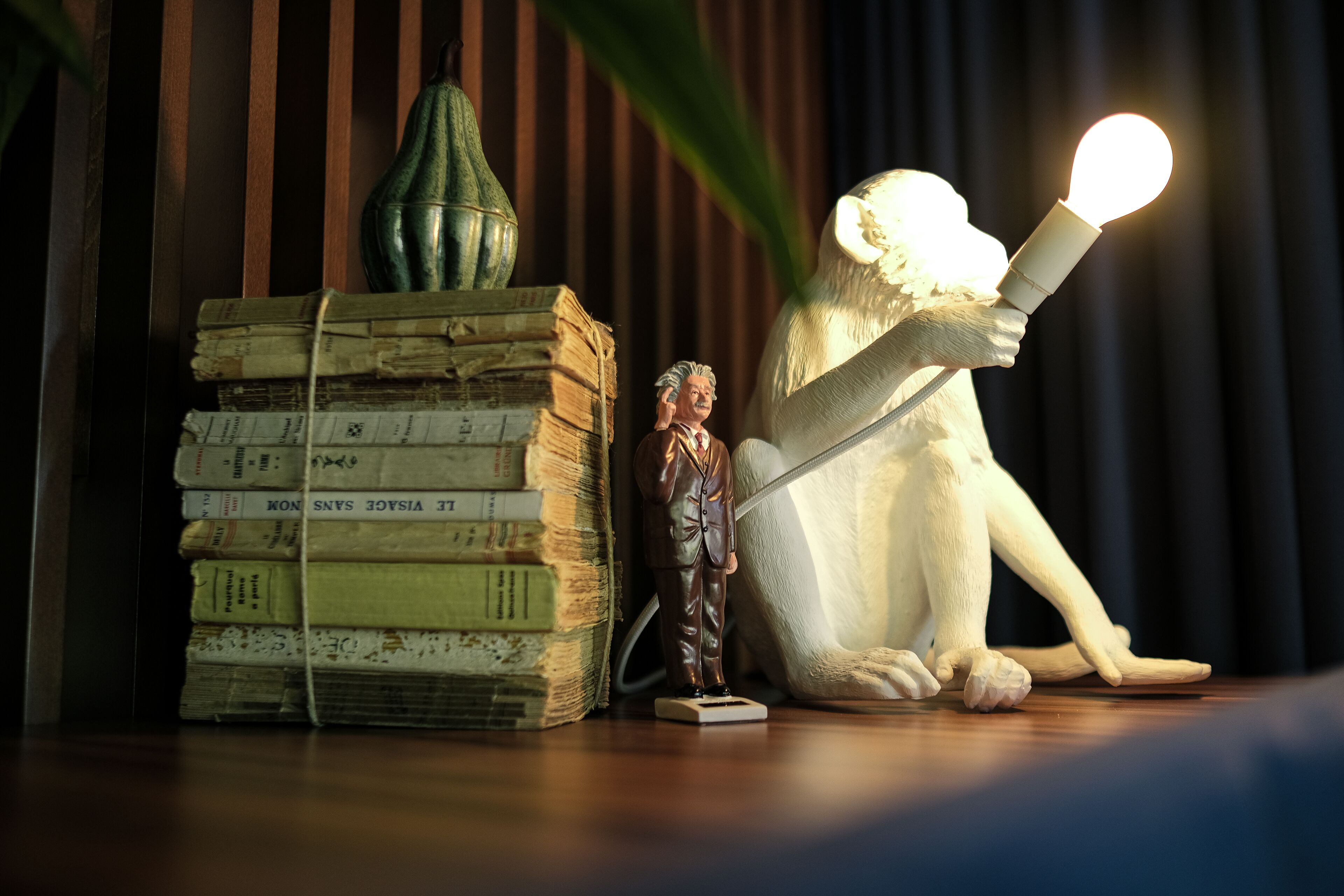 Una estatua de un hombre leyendo un libro, una pila de libros y un jarrón verde sobre una superficie de madera.