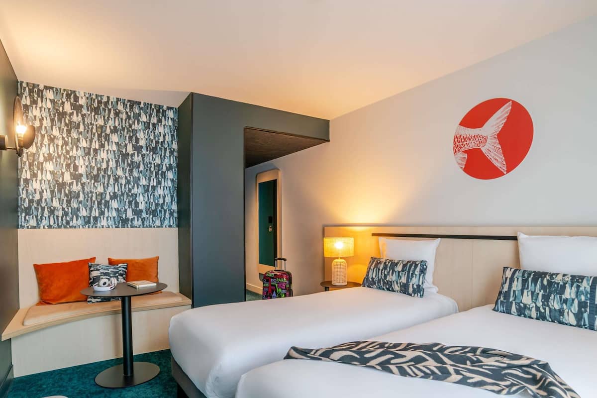 A hotel room with two beds, a desk, a chair, a wall with a red circular design, and a patterned wall.