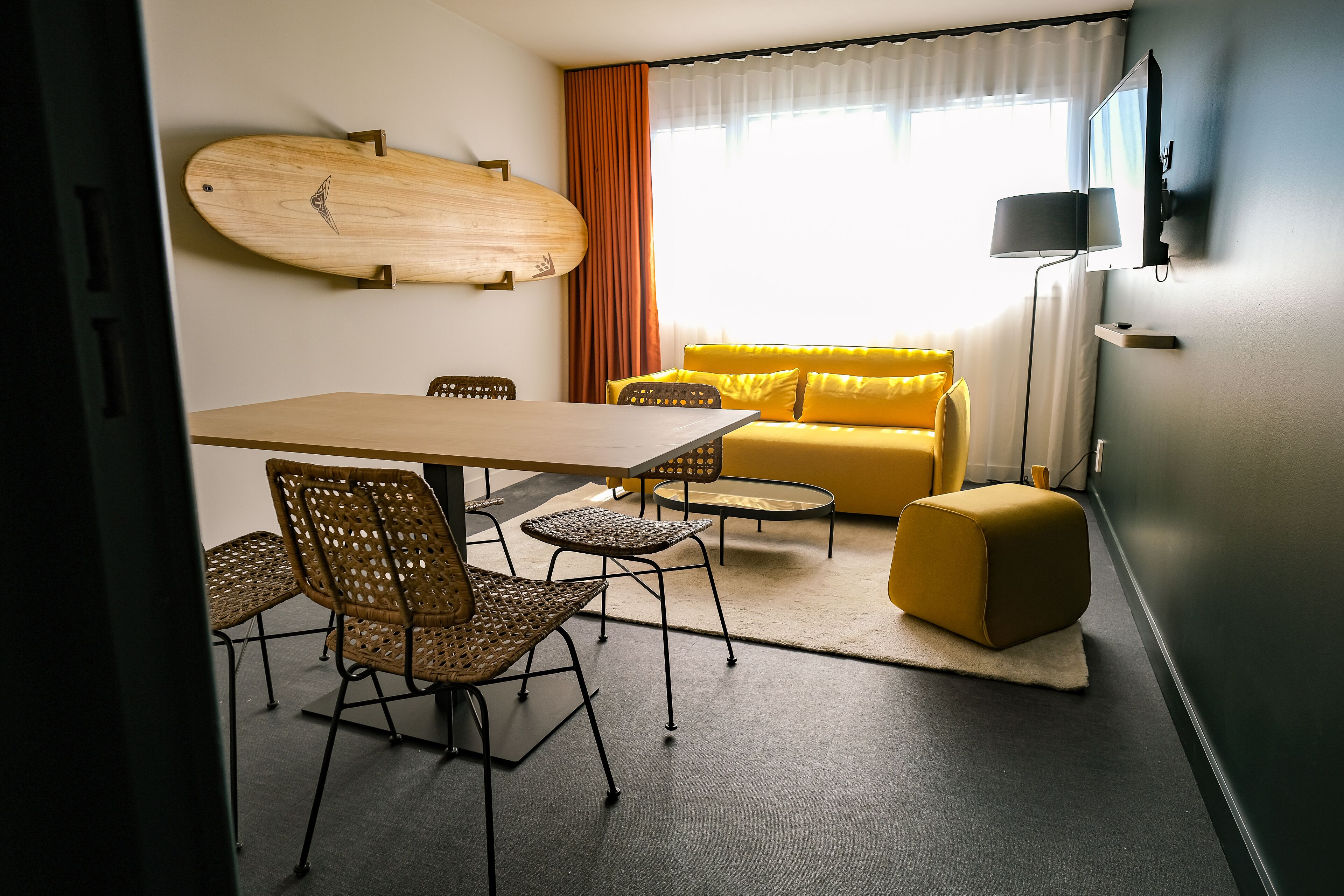 A modern living room with a dining area, a yellow sofa, wicker chairs, and a surfboard on the wall.