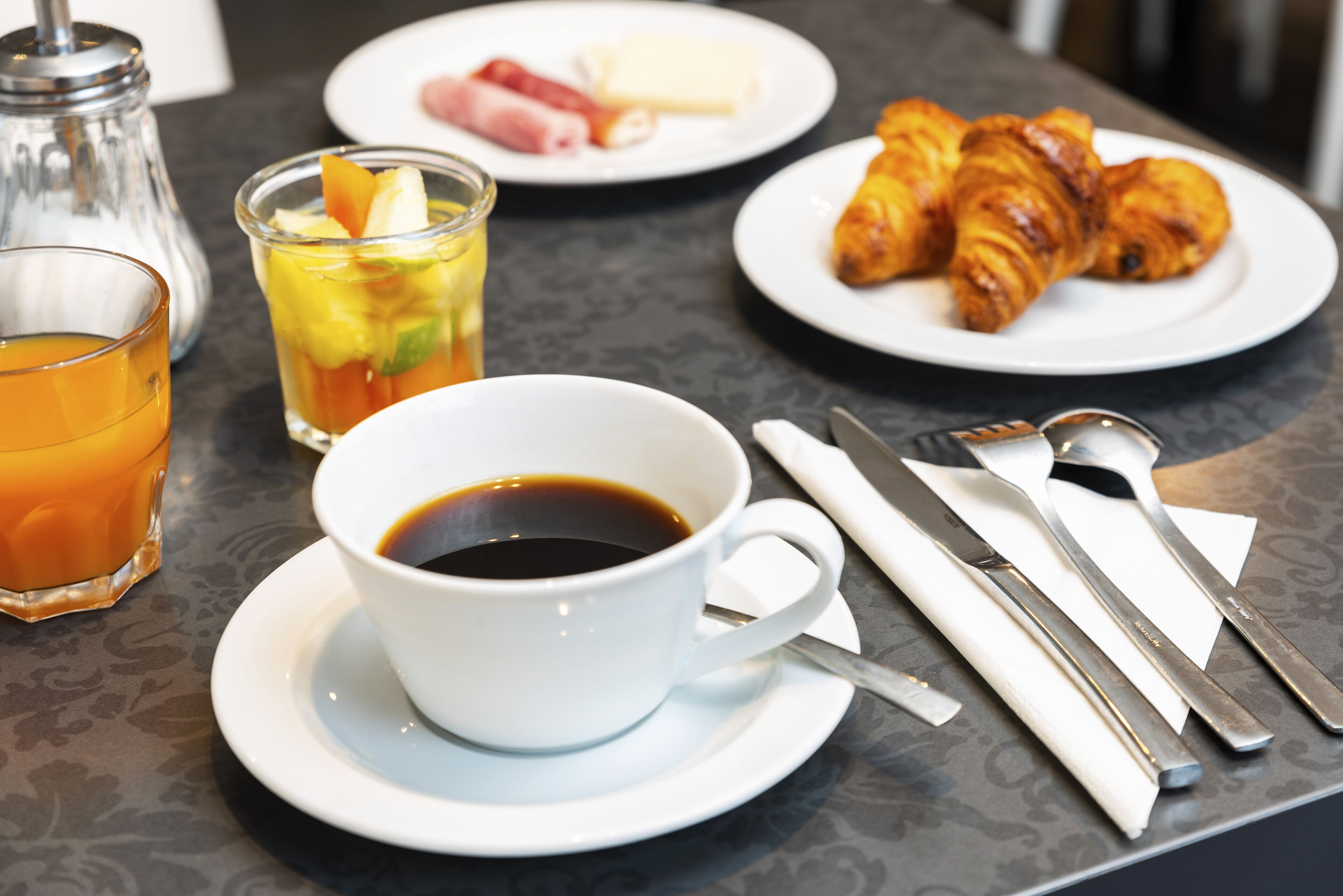 Una mesa con una taza de café, un vaso de zumo de naranja, croissants y un plato de jamón y queso.