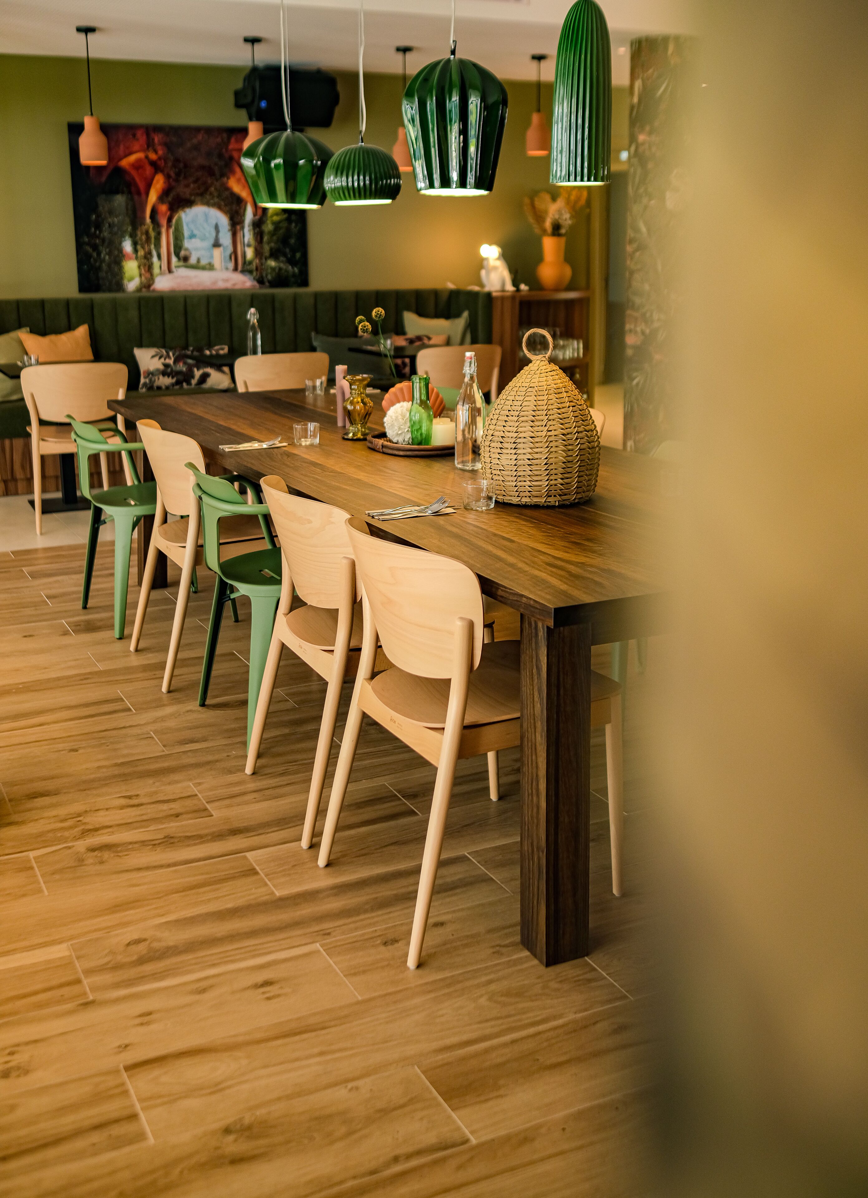 A dining area with a wooden table, green and beige chairs, hanging green pendant lights, and a woven basket on the table.