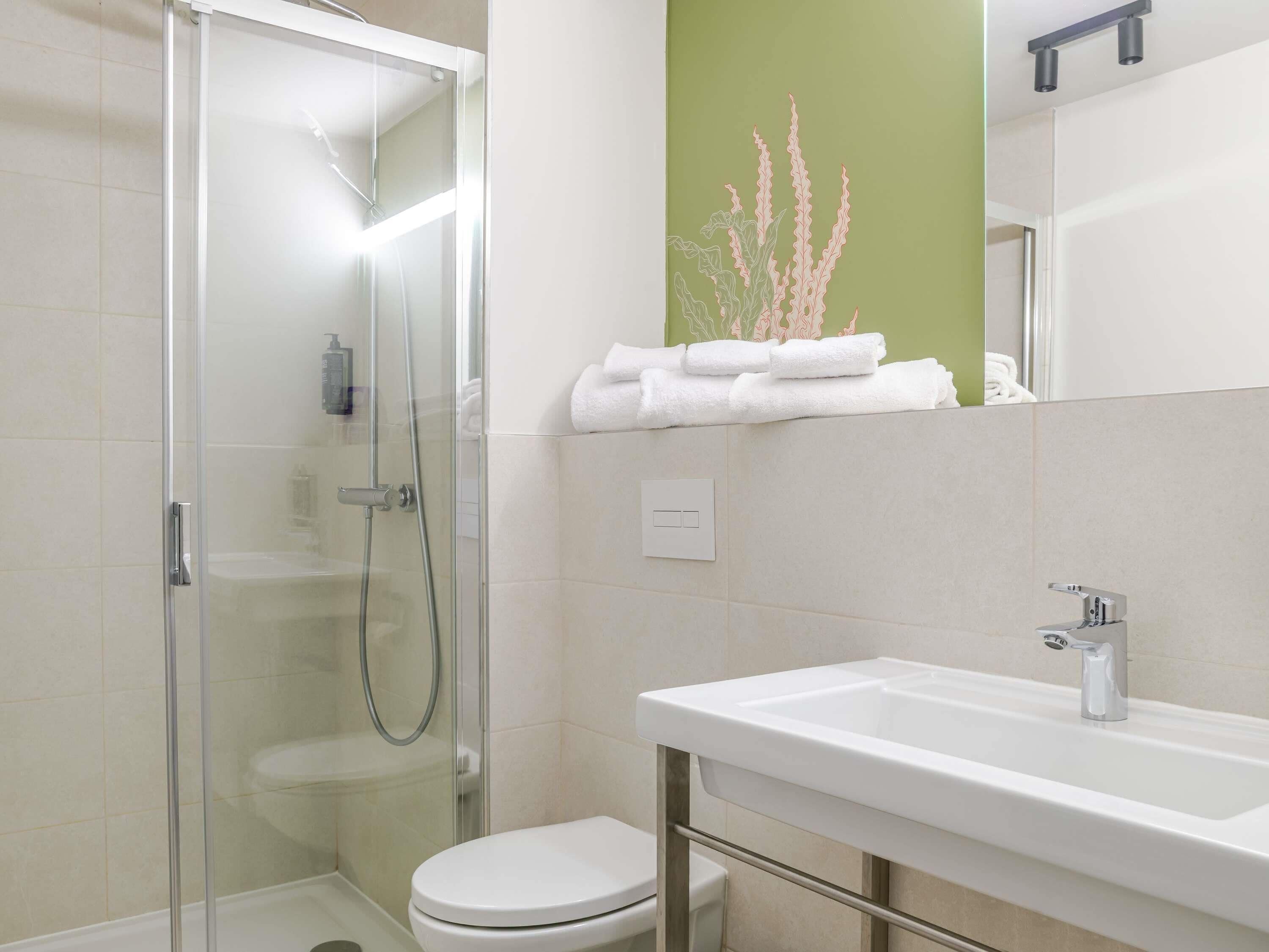 A modern bathroom with a glass shower enclosure, a white toilet, a sink with a chrome faucet, and a green accent wall.