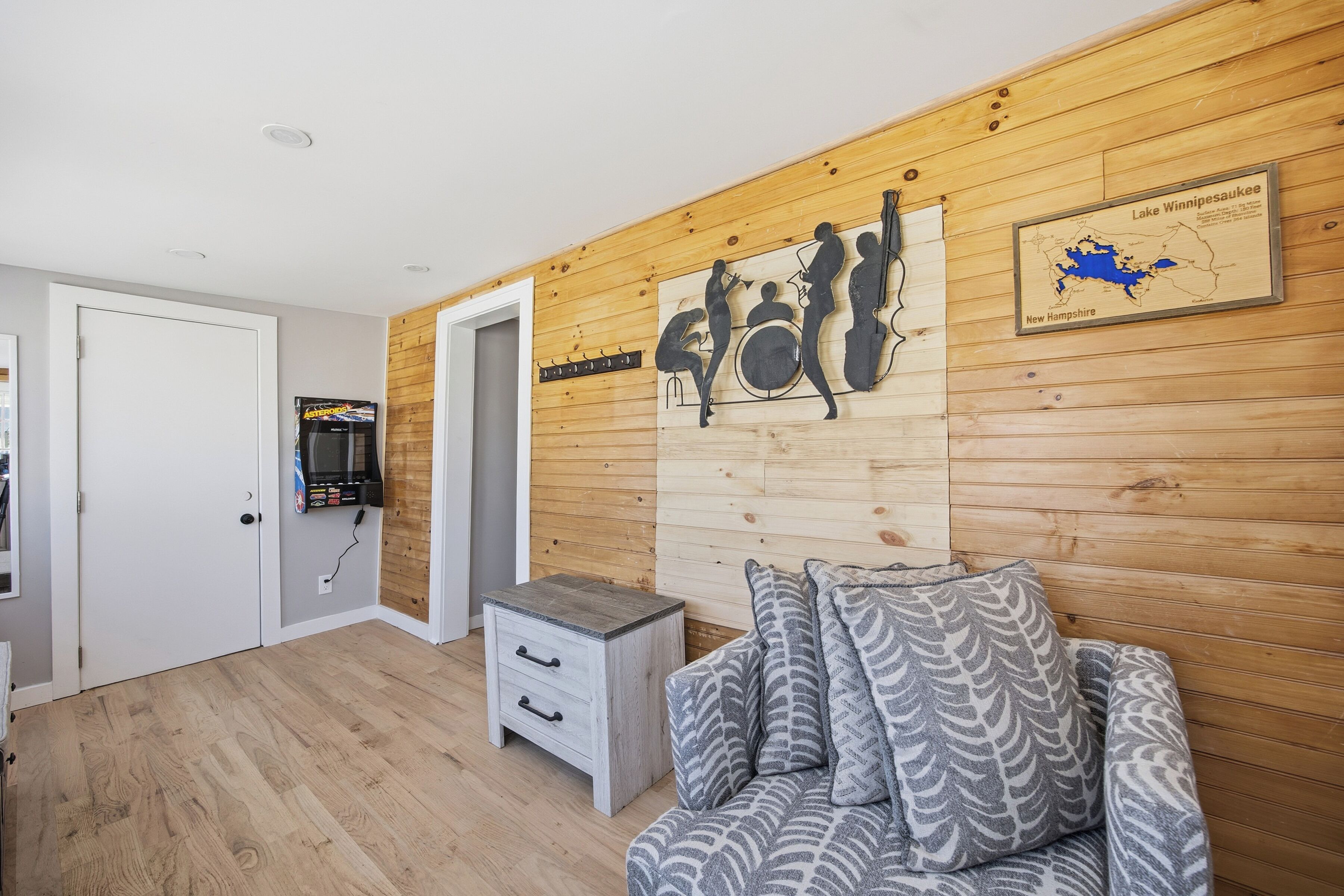Ein Zimmer mit Holzwandverkleidung, einer weißen Tür, einem grauen Sofa mit Muster und einem kleinen weißen Nachttisch. Ein an der Wand hängendes Kunstwerk zeigt eine Szene mit musikalischen Elementen.