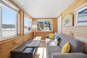 Una cabaña con interior de madera, un sofá gris con cojines amarillos, un banco de madera y una ventana con vistas a árboles y un paisaje nevado.