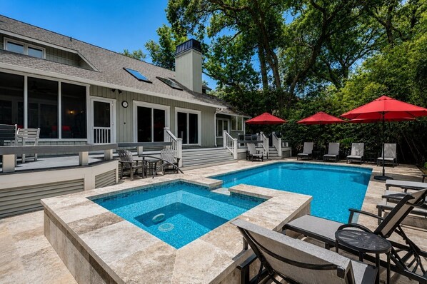 Une piscine dotée d’une partie plus profonde, entourée de chaises longues et de parasols rouges, avec un patio couvert et une maison en arrière-plan.