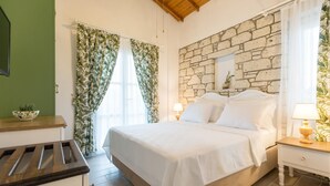 A bedroom with a stone wall, a bed with white linens, a wooden nightstand with a lamp, and a window with curtains. - Melekaki Alacati (Cesme)