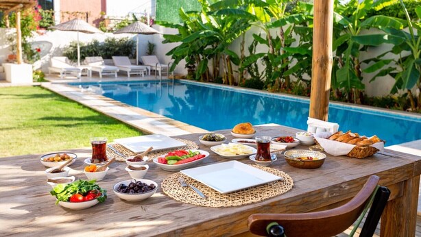A poolside dining table with various dishes and drinks, surrounded by lush greenery and a pool. - Melekaki Alacati (Cesme)