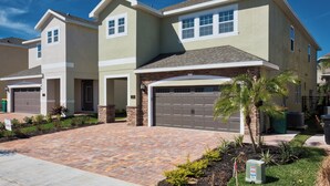 A two-story house with a brick driveway, a garage, and a small garden with palm trees.