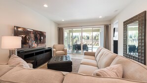 A spacious living room with a large flat-screen TV, a beige sofa, a black ottoman, and a dining area with a table and chairs.