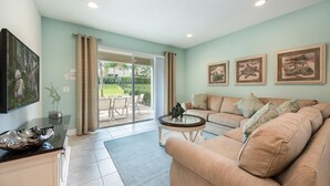 A living room with a beige sofa, a glass coffee table, and a flat-screen TV on a cabinet. There are framed pictures on the wall and a sliding glass door leading to a patio.