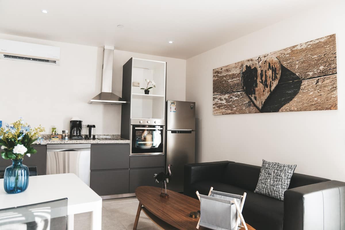 A modern kitchen with a built-in oven and refrigerator, a dining area with a wooden table, and a black sofa with a patterned pillow.