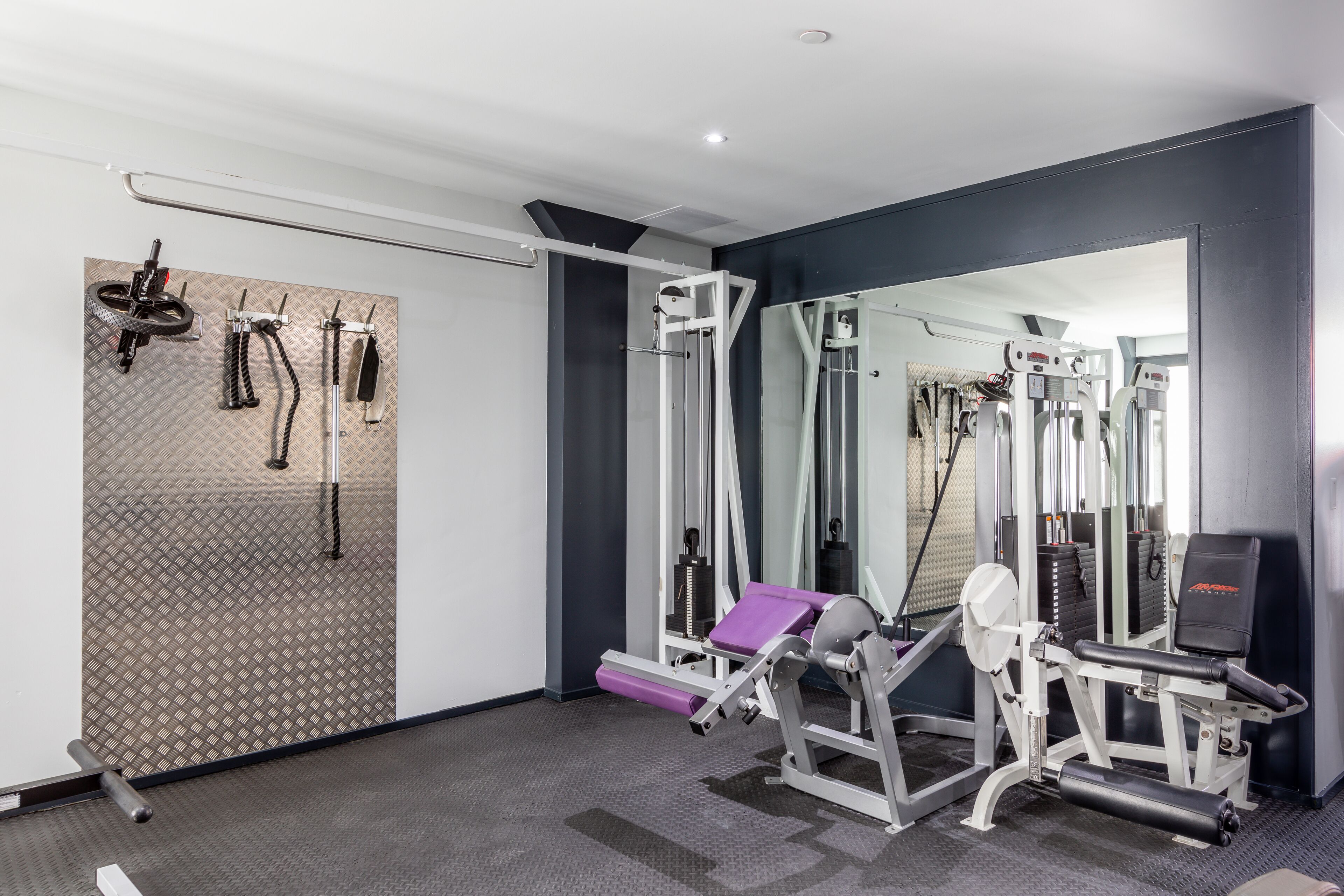 Ein Fitnessstudio mit verschiedenen Trainingsgeräten, einer Spiegelwand und einem gemusterten Boden.